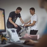 Ein Mann sitzt auf einem Ergometer. Ein zweiter Mann steht daneben und zeigt ihm etwas.