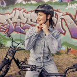 Eine Frau steht mit einem E-Bike vor einer Wand mit Graffiti und hantiert am Kinnriemen ihres Fahrradhelms.