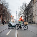 Eine Frau fährt auf einem E-Lastenrad mit Kinderanhänger quer über eine Straße. Im Hintergrund ist der Berliner Fernsehturm zu erkennen.