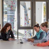 Fünf Frauen sitzen an einem Tisch und schauen Dokumente an. Hinter ihnen sind durch Fenster Kletterwände zu sehen.