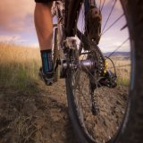 Blick über den Hinterreifen eines Mountainbikes auf die Waden der fahrenden Person.