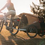 Eine Person fährt mit einem E-Bike und Fahrradanhänger mit einem Hund darin über eine Wiese.