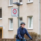Ein Mann fährt auf einem Fahrrad auf einer Straße. Im Hintergrund ist vor einem Wohnhaus ein Verkehrsschild mit Geschwindigkeitsbegrenzung 30 km/h zu sehen.