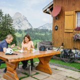 Zwei Personen an einem Tisch vor einer Berghütte. Zwei E-Bikes lehnen an der Wand.
