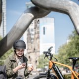 Eine Frau sitzt neben einem abgestellten E-Bike auf einer Bank und schaut lachend auf ein Smartphone.