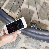Eine Person hält ein Smartphone vor einen Fahrradreifen, an dessen Ventil ein kleines, schwarzes Gerät montiert ist.