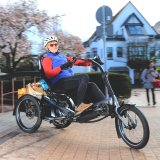 Eine Person auf einem Dreirad mit Tretkurbel, Motorunterstützung und Gepäck zwischen den beiden Hinterrädern.