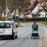 Ein weißes Auto überholt eine Person auf einem Fahrrad mit Anhänger auf einer Straße ohne Fahrradspur in einem Wohngebiet.