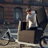 Ein Mann schaut in die Ladebox eines Lastenrades. Den Deckel der großen Box, die sich zwischen Lenker und Vorderrad befindet, hält er mit einer Hand offen.