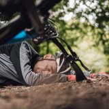 Eine Person mit Fahrradhelm liegt im Wald auf dem Boden. Im Vordergrund sind Teile eines Fahrrads zu erkennen.