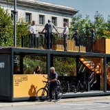 In einer teils offenen Struktur am Straßenrand, die einer Bushaltestelle ähnelt, stellen Menschen Fahrräder ab. Eine Treppe führt auf das Dach, wo Menschen an einem Geländer stehen.