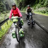 Zwei Personen fahren auf einer nassen Straße in einem Tal im Wald auf den Betrachter zu.