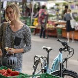 Eine Frau steht mit Fahrradhelm in der Hand an einem Gemüsestand auf einem Markt. Neben ihr steht ein E-Bike.