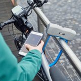Blick über die Schulter einer Person, die ein Smartphone vor eine Bügelschloss hält, mit dem ein E-Bike an ein Geländer angeschlossen ist.