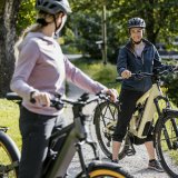 Zwei Frauen mit E-Bike stehen mit E-Bikes auf einem Radweg und lächeln einander zu.