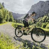 Eine Frau fährt auf einem E-Bike einen Schotterweg im Gebirge entlang.
