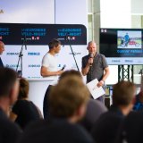 Drei Männer stehen auf einer Bühne vor Publikum. Einer spricht in ein Mikrophon.