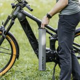Eine Person steht neben einem E-Bike und hält den länglichen Akku in der Hand, der im Rahmen montiert wird.