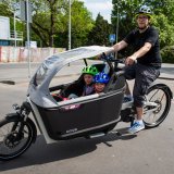 Ein Mann fährt ein Lastenrad mit zwei Kindern und Regenverdeck.