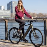 Eine Frau sitzt auf einem E-Bike und hält sich am Geländer eines Flussufers fest.