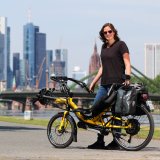 Eine Frau steht mit einem gelben Liegerad mit Versicherungskennzeichen am Gepäckträger vor der Skyline einer Großstadt.