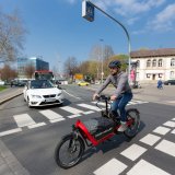 Ein Lastenradfahrer überquert eine Straße innerhalb einer Fahrbahnmarkierung. Ein Auto wartet im Hintergrund.