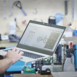 Eine Person hält in einer Werkstatt einen Laptop vor sich, auf dessen Bildschirm ein E-Bike zu sehen ist.