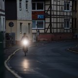Ein Radfahrer fährt in der Dämmerung eine leere Straße zwischen Häusern entlang. Das Licht am Fahrrad leuchtet deutlich in die Kamera.