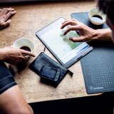 Ein Tablet liegt zwischen zwei Kaffeetassen und einem Navigationsgerät für Fahrräder auf einem Tisch. Zwei Personen beschäftigen sich mit einer Kartenanwendung darauf.