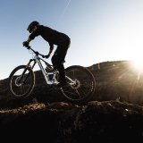 Eine Person fährt in offener Gebirgslandschaft vor der tiefstehenden Sonne ein E-Mountainbike.