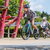 Ein Junge fährt neben einem Klettergerüst Fahrrad. Im Hintergrund folgt ein zweites Kind.