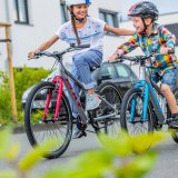 Ein Mädchen und ein kleinerer Junge fahren auf Fahrrädern. Das Mädchen lächelt dem Jungen zu und hat eine Hand auf seiner Schulter, um ihn zu unterstützen.