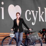 Eine Frau steht an einem Fahrrad gelehnt mit einem Helm am Lenker hängend. Im Hintergrund sind Fahrradständer zu erkennen und ein großes weißes Herz an einer grauen Wand mit dem Wort ?cykler? daneben.