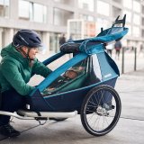 Eine Frau hockt vor einem Kinderanhänger an einem Fahrrad und greift hinein.