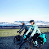 Eine Frau und ein Mann fahren lächelnd mit bepackten Fahrrädern vor einer entfernten Bergkulisse in offener Landschaft entlang.