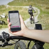 Eine Person hät ein Smartphone mit Karten-App darauf über den Lenker eines E-Bikes. Im Hintergrund fährt eine Person einen Wirtschaftsweg entlang.