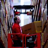 Ein Mann steht mit einem Paket in den Händen auf einem Fahrzeug zwischen zwei Hochregalen in einem Lager. 