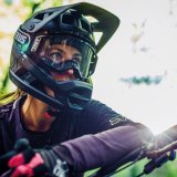 Frau mit Fullface-Helm mit Mountainbike im Wald bei Sonnenschein.