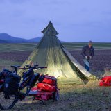 Am Rand eines Feldes steht ein Pyramidenzelt. Dahinter geht ein Mann entlang, im Vordergrund steht ein Lastenrad.