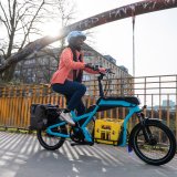 Eine Frau fährt auf einem blauen Lastenrad mit einem gelben Getränkekasten auf der Ladefläche über eine Brücke in der Stadt.