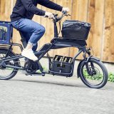Mensch fährt beladenes Cargobike vor Holzwand.