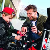 Ein Mann und ein Junge hocken neben einem Kinderrad. Der Junge hat ein Werkzeug in der Hand.