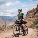 Ein Mann steht mit einem Fahrrad mit Bikepacking-Taschen auf einem Schotterweg in einer trockenen Gebirgslandschaft.
