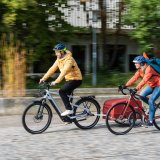 Ein Mann auf einem E-Bike mit Anhänger und eine Frau auf einem Rennrad fahren nebeneinander einen gepflasterten Weg entlang.