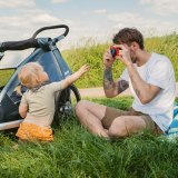 Ein Mann sitzt auf einer Wiese neben einem Fahrradanhänger und fotografiert mit einer bunten Kamera ein kleines Kind.