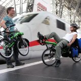 Ein Mann mit einem zusammengeklappten Liegerad in der Hand und eine Frau auf einem Liegerad mit Taschen daran auf einem Bahnsteig vor einem weiß-roten Zug.