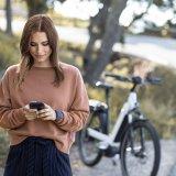 Eine Frau steht am Straßenrand vor einem Fahrrad und schaut auf ihr Smartphone.