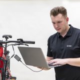 Ein Mann steht neben einem E-Bike in einem Montageständer. In der Hand hält er einen Laptop, der über Kabeln mit dem Rad verbunden ist.
