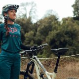 Eine Frau steht vor einem Zaun vor Bäumen und hält mit der linken Hand ein Mountainbike am Lenker.