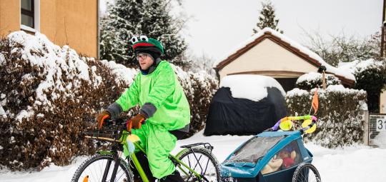 Ein Mann in einem Frosch-Kostüm fährt auf einem Fahrrad mit Kinderanhänger auf einer schneebedeckten Fahrbahn.
